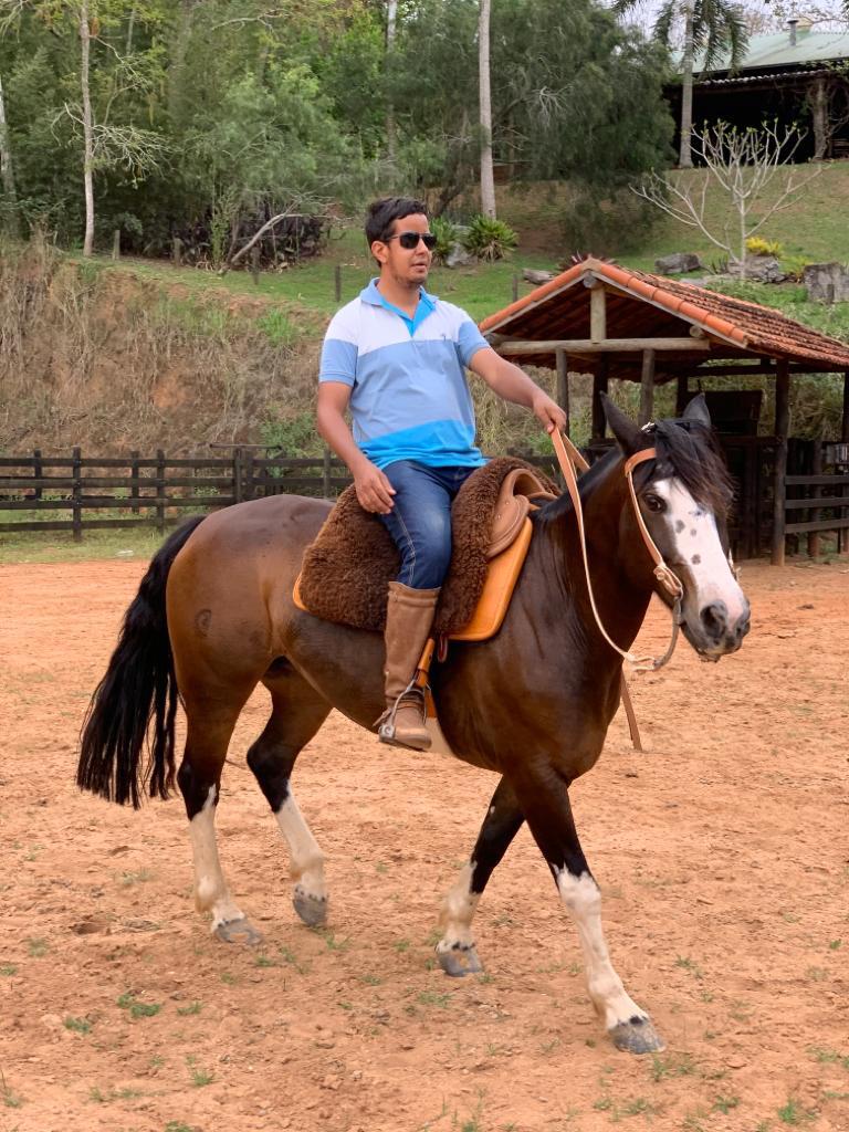 Na imagem Hugo Antônio está montado em um cavalo de cor marrom. Ele está vestido com uma uma blusa de manga curta, na cor azul, uma calça jeans azul e calçado com uma bota. 
